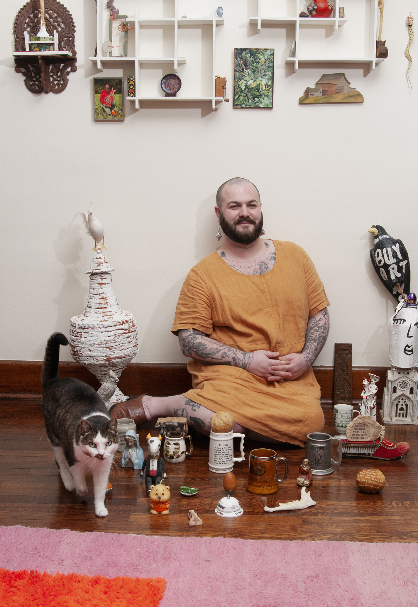A bearded person sitting of the floor of their room surrounded by objects and shelves above, and a tabby cat.